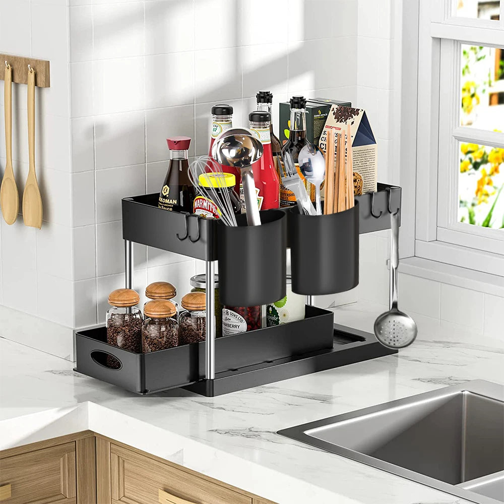 Zwart uitschuifbaar keukenrek met twee niveaus, zijhaken en extra opbergbekers, gevuld met kruidenpotjes, kookgerei en diverse keukenbenodigdheden, geplaatst op een aanrecht naast een spoelbak in een moderne keuken.