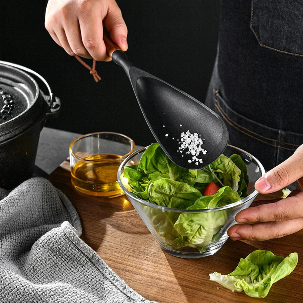 Zwarte siliconen opscheplepel met houten handvat waarmee zout wordt toegevoegd aan een frisse salade in een glazen kom, met olijfolie en keukentextiel op een houten keukenblad. Ideaal voor stijlvol koken en serveren.