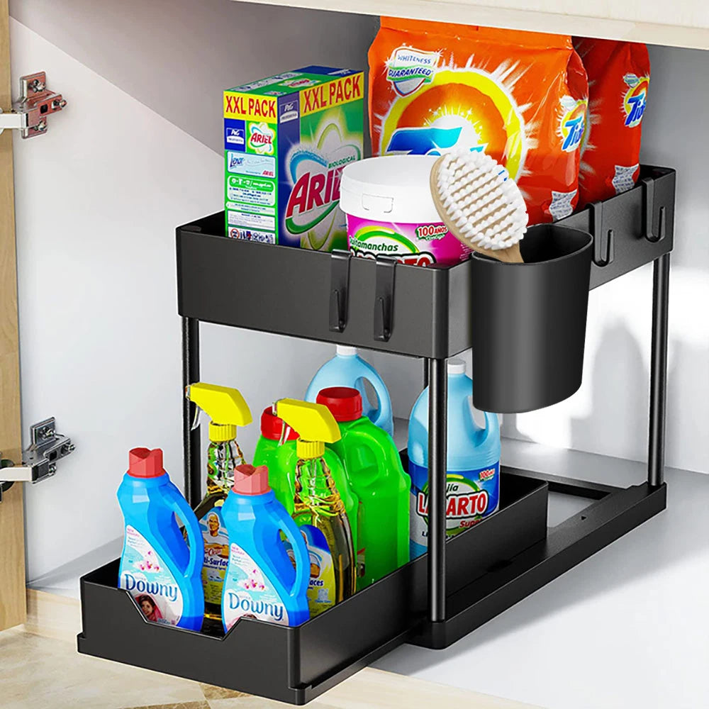 Uitschuifbaar opbergrek voor onder de gootsteen met twee niveaus, inclusief zijhaken en een extra opbergvak, gevuld met diverse schoonmaakmiddelen en wasmiddelen zoals Ariel en Downy, ideaal voor het efficiënt organiseren van schoonmaakproducten in keukenkast of badkamer.