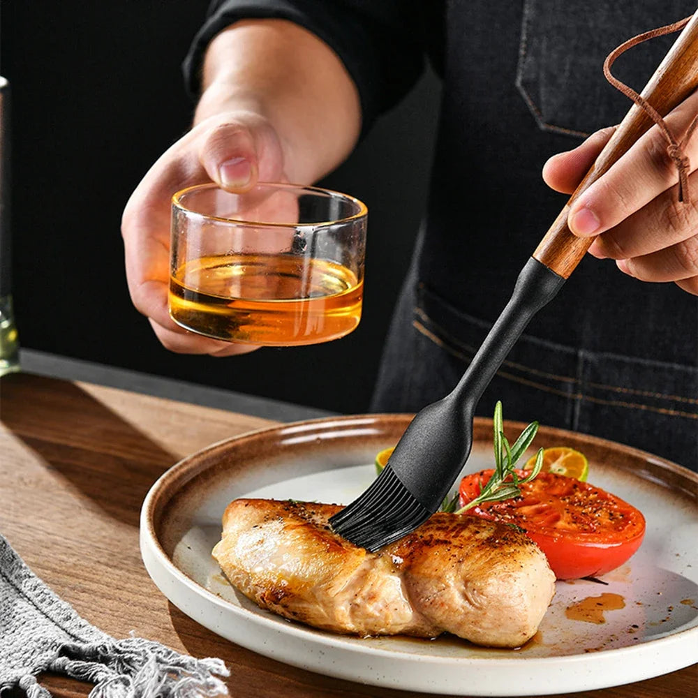 Zwarte siliconen keukenkwast met houten handvat, gebruikt om marinade aan te brengen op een gegrild stuk kipfilet, geserveerd met tomaat en kruiden op een keramisch bord, met een glaasje marinade op de achtergrond. Ideaal voor het bereiden en serveren van smaakvolle gerechten.