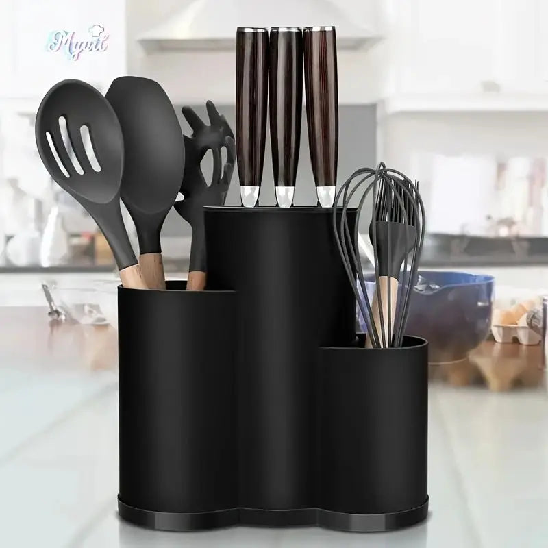 Zwarte keukenorganizer met drie compartimenten gevuld met messen, kunststof spatels en een garde, geplaatst op een keukenblad met een moderne keuken op de achtergrond.