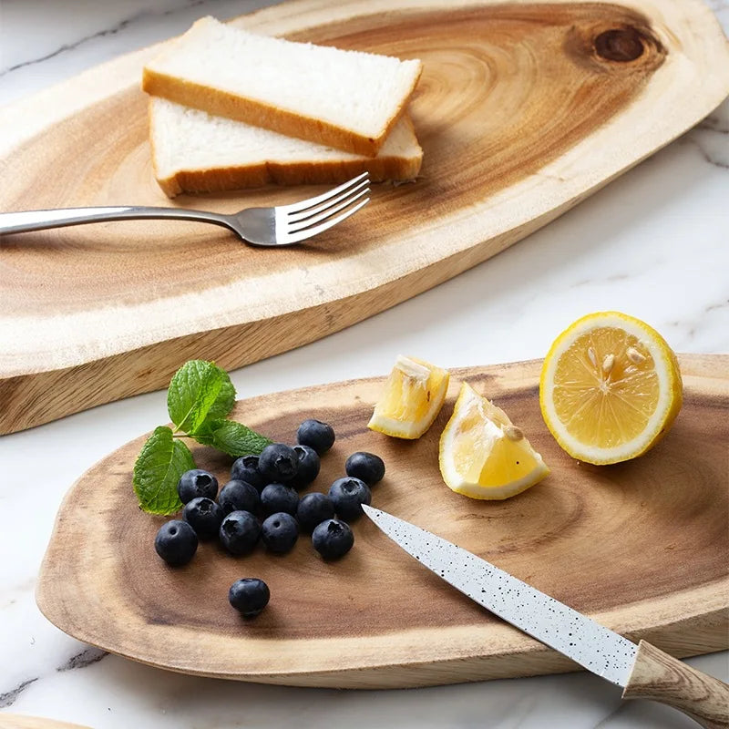 "Twee ovale acaciahouten snij- en serveerplanken met citroen, blauwe bessen, munt, brood en een mes op een marmeren aanrecht.