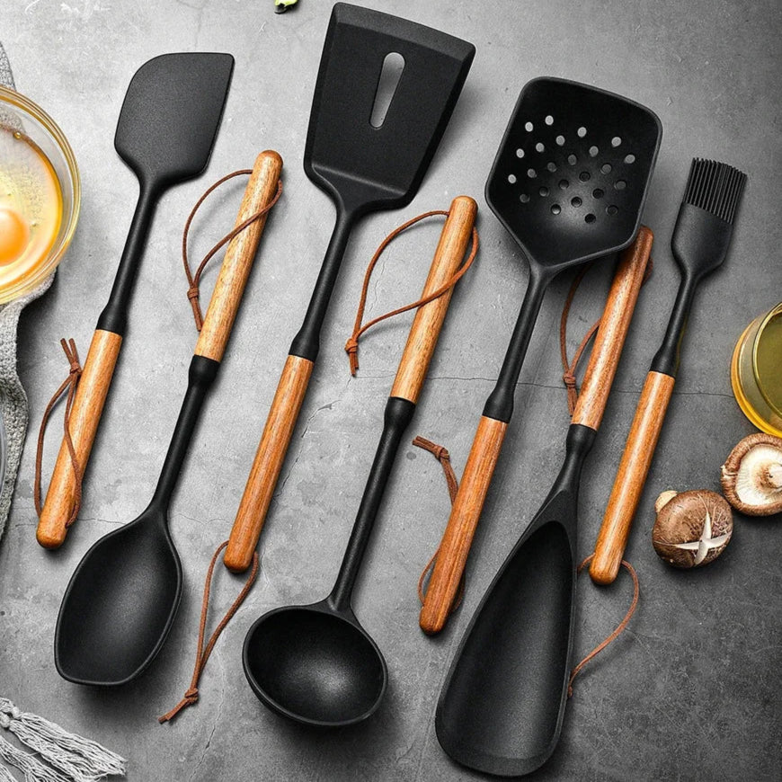 Complete set zwarte siliconen keukenhulpen met houten handvatten, inclusief spatel, soeplepel, bakspaan, schuimspaan, opscheplepel en keukenkwast, stijlvol gepresenteerd op een grijze stenen ondergrond met ingrediënten rondom. Perfect voor koken en bakken.